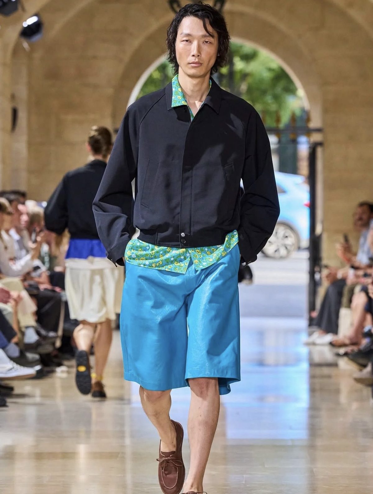 Modelo con shorts de piel turquesa, camisa estampada verde y chaqueta negra en un desfile de primavera 2027