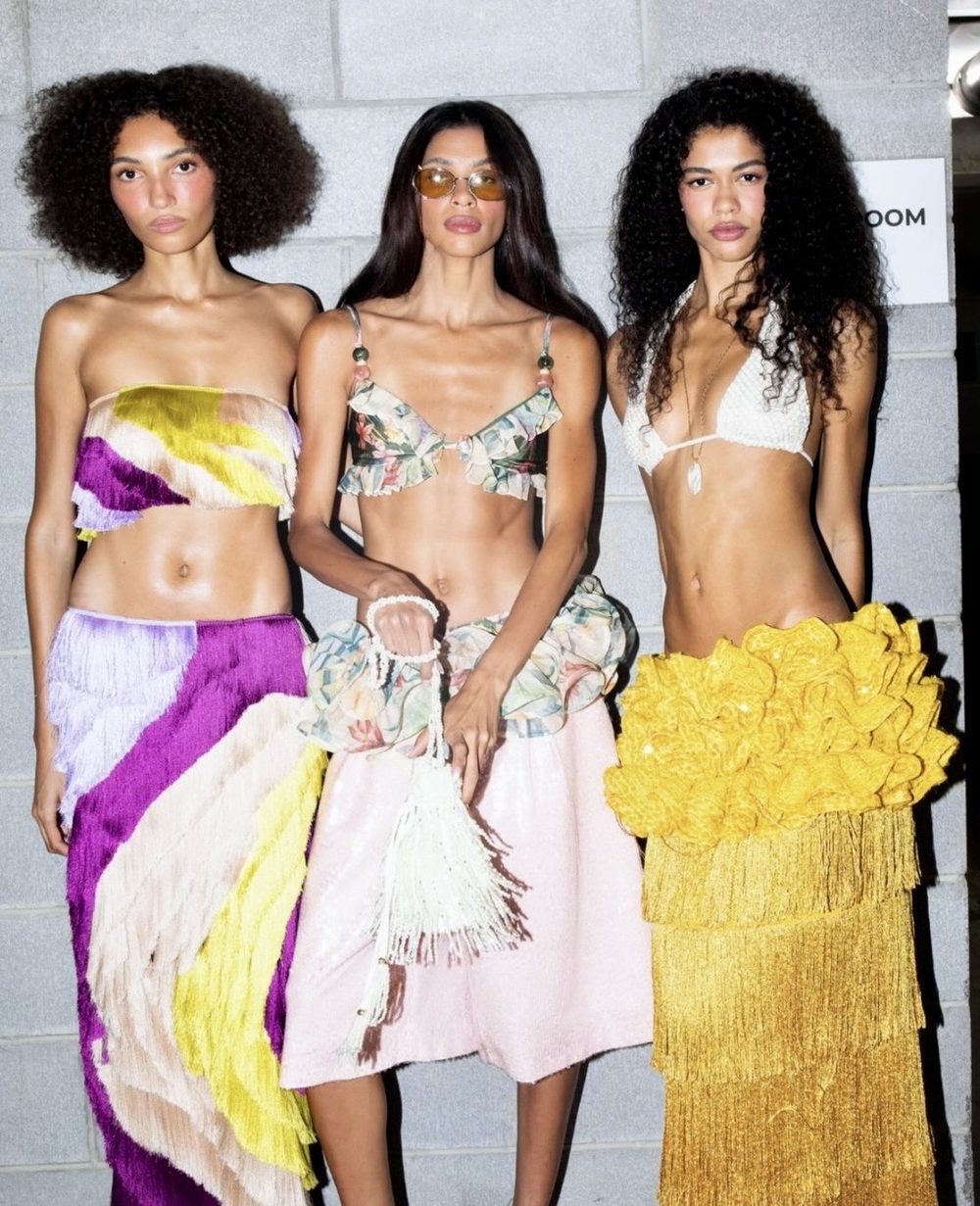 Tres modelos en backstage con tops cortos y faldas de flecos en morado, rosado y amarillo
